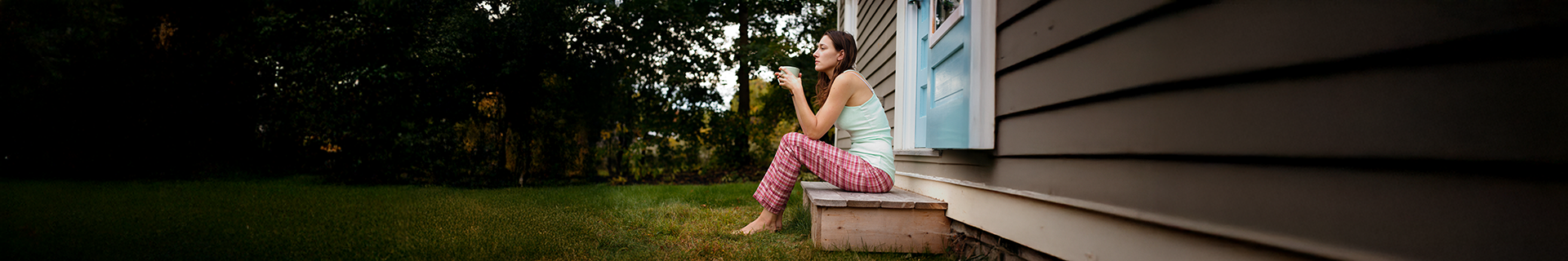 A woman sitting in her pyjamas while drinking coffee on the steps outside her house while barefoot with her feet in the grass
